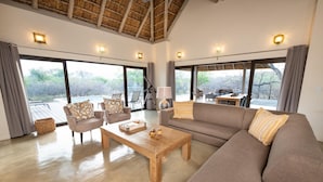 A spacious living area with a large sofa, two armchairs, a wooden coffee table, and a dining table. There are large windows providing views of the outdoors.