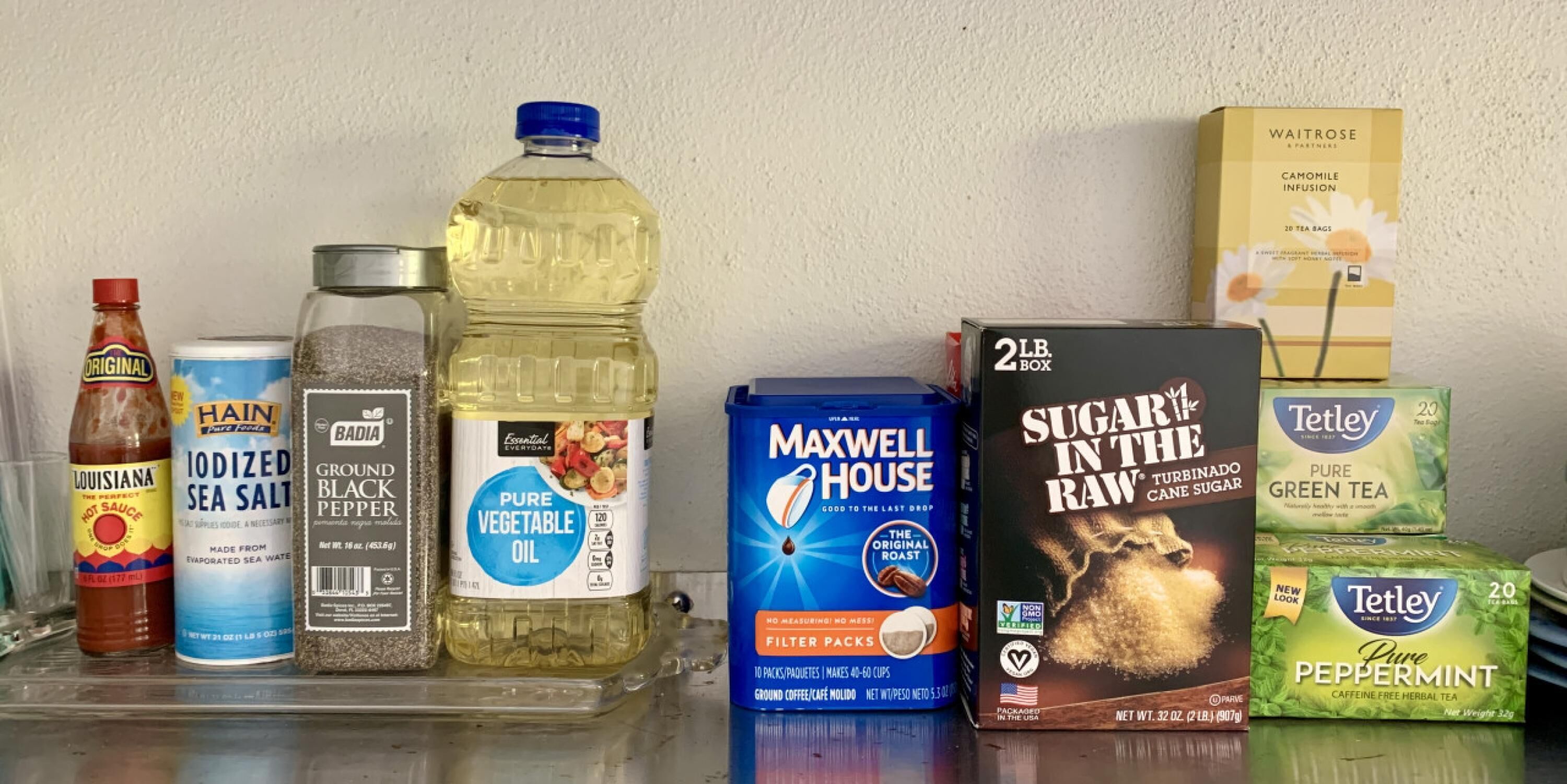 A shelf with various food and drink items including a bottle of hot sauce, iodized sea salt, vegetable oil, ground black pepper, Maxwell House coffee, sugar, raw cane sugar, and tea bags.