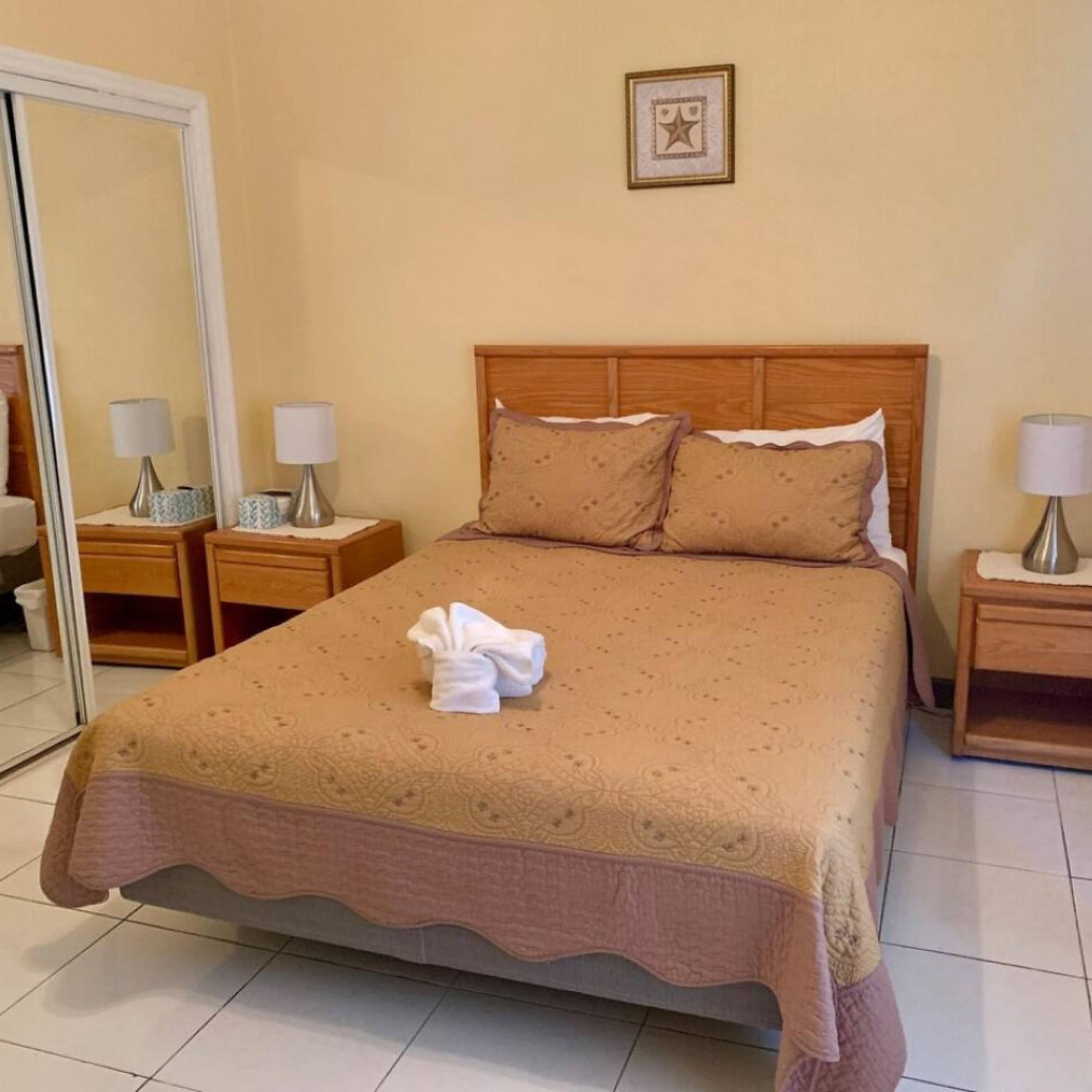 A neatly made bed with a patterned bedspread, two pillows, and a folded towel on the bed. There are two bedside tables with lamps and a mirror on the left side.