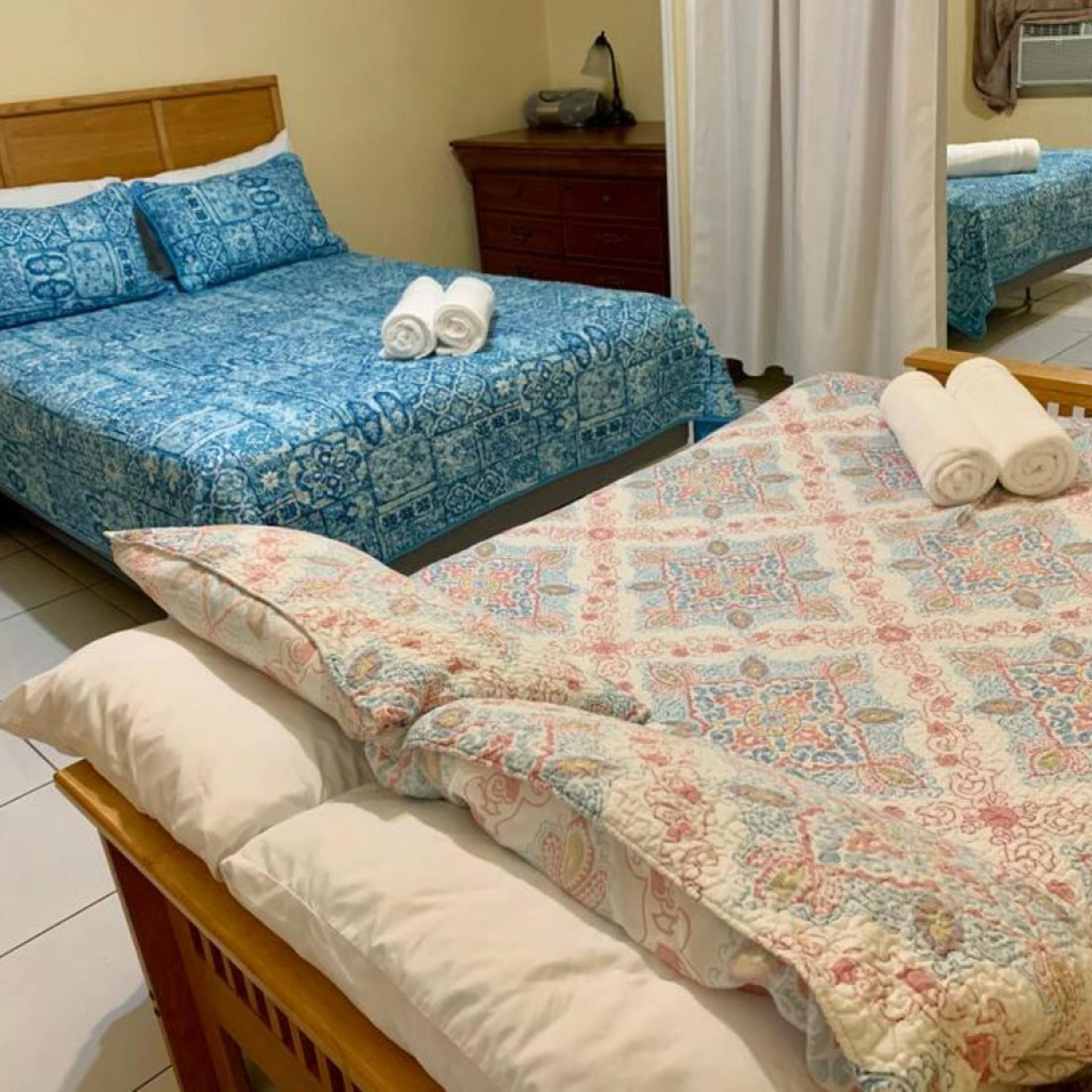 A room with two beds, one with blue patterned bedding and the other with pink and white patterned bedding. There are two rolled towels on each bed.