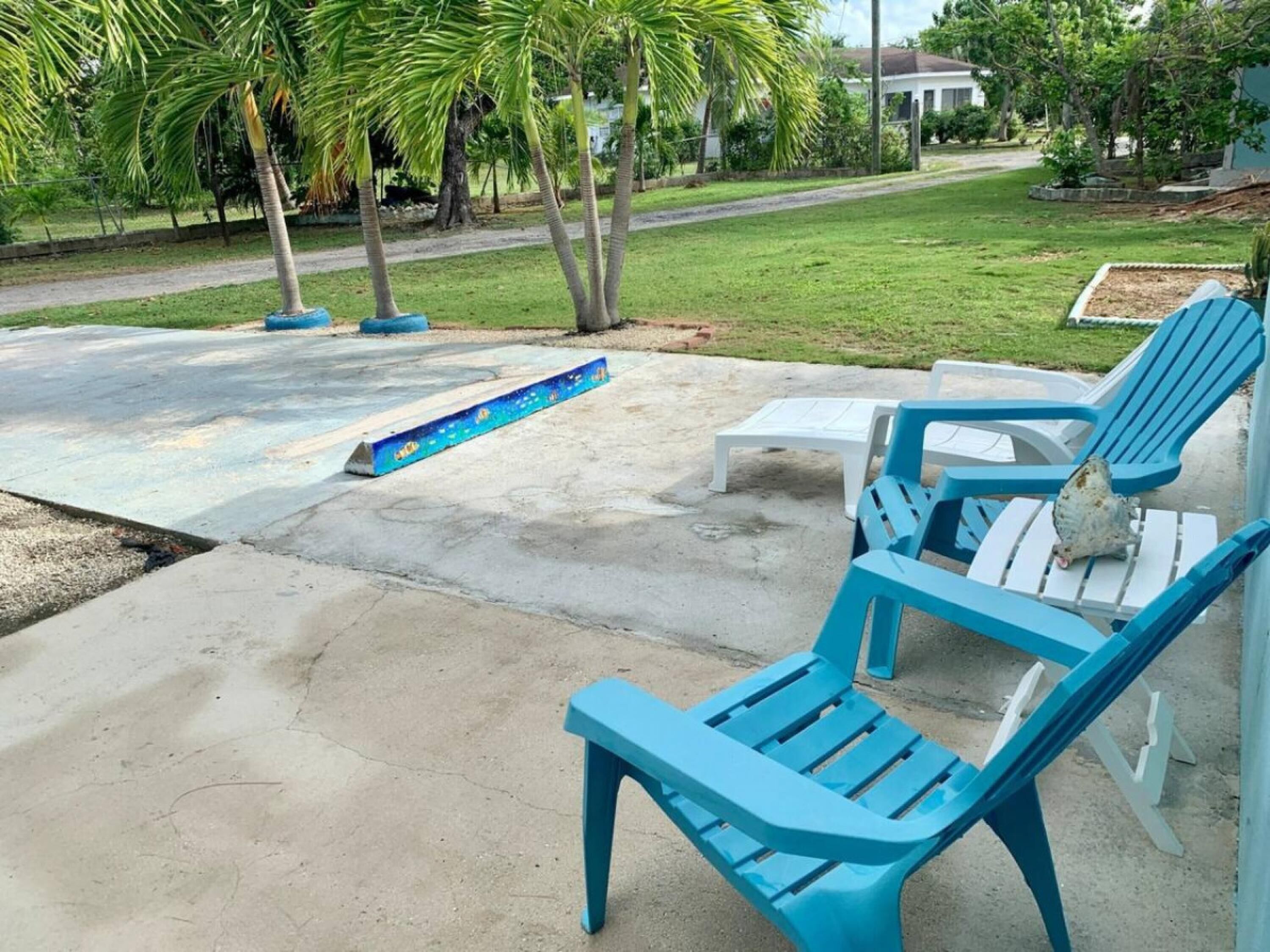 A concrete patio with blue Adirondack chairs and a small table, surrounded by palm trees and a grassy lawn.