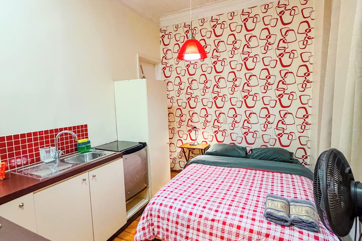 A compact room with a kitchenette, a bed with a checkered bedspread, a patterned wall, and a ceiling light fixture.