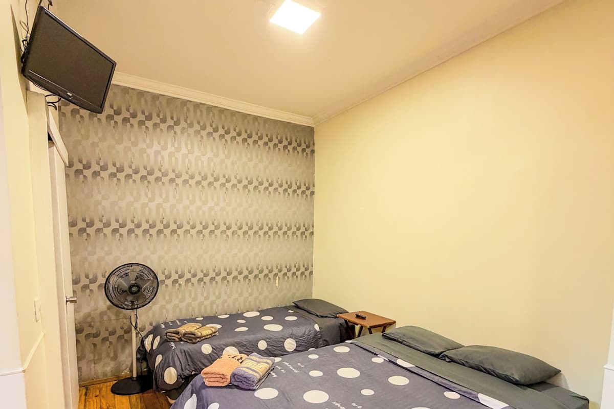 A room with two beds, a standing fan, a television mounted on the wall, and a patterned wallpaper.