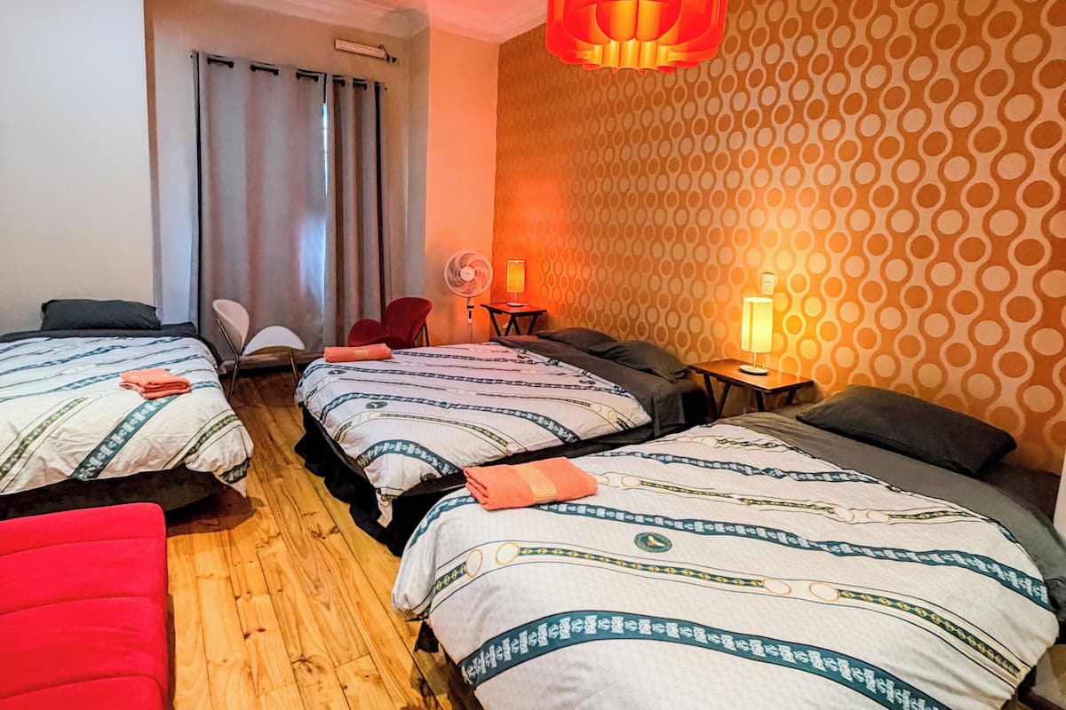 A hotel room with two beds, a red chair, a wooden floor, a patterned wall, and a hanging light fixture.