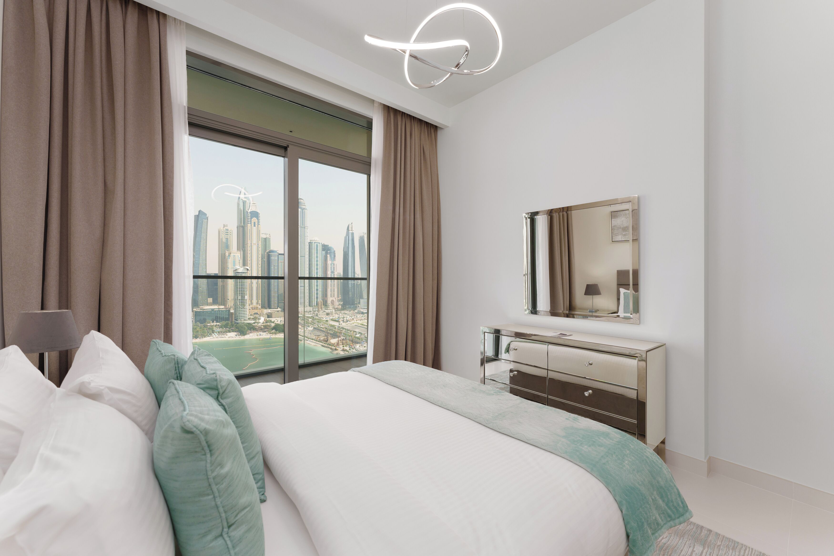 Ein modernes Hotelzimmer mit einem großen Bett, einem Schminktisch, einem Spiegel und Blick auf eine Stadtlandschaft durch ein großes Fenster.
