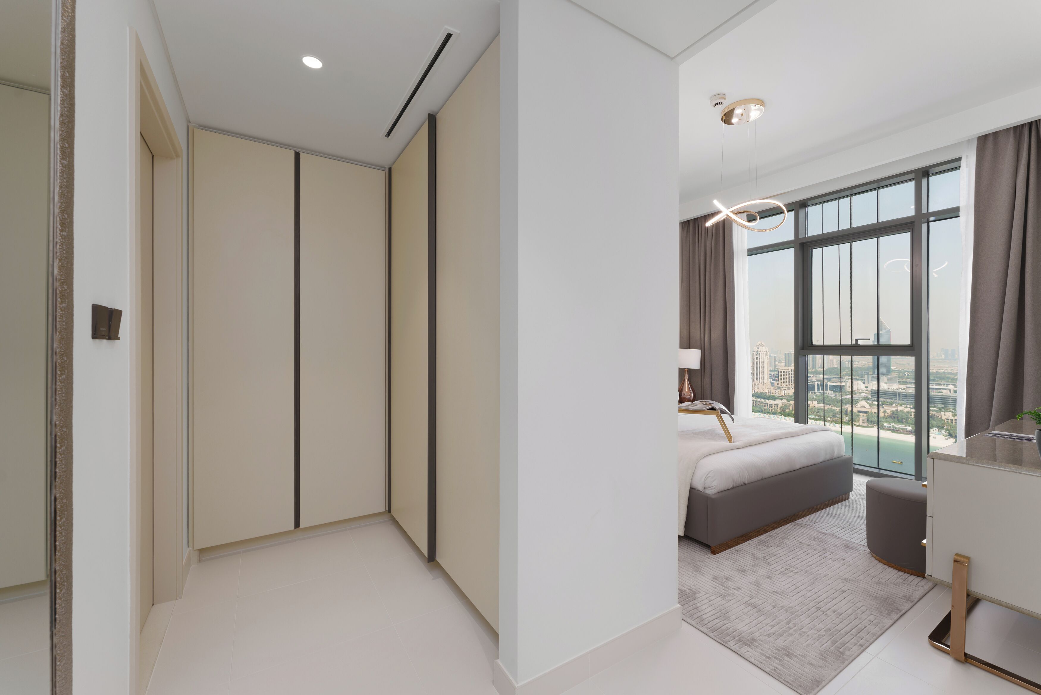 A modern bedroom with a large bed, a walk-in closet, and a view of a cityscape through a large window.