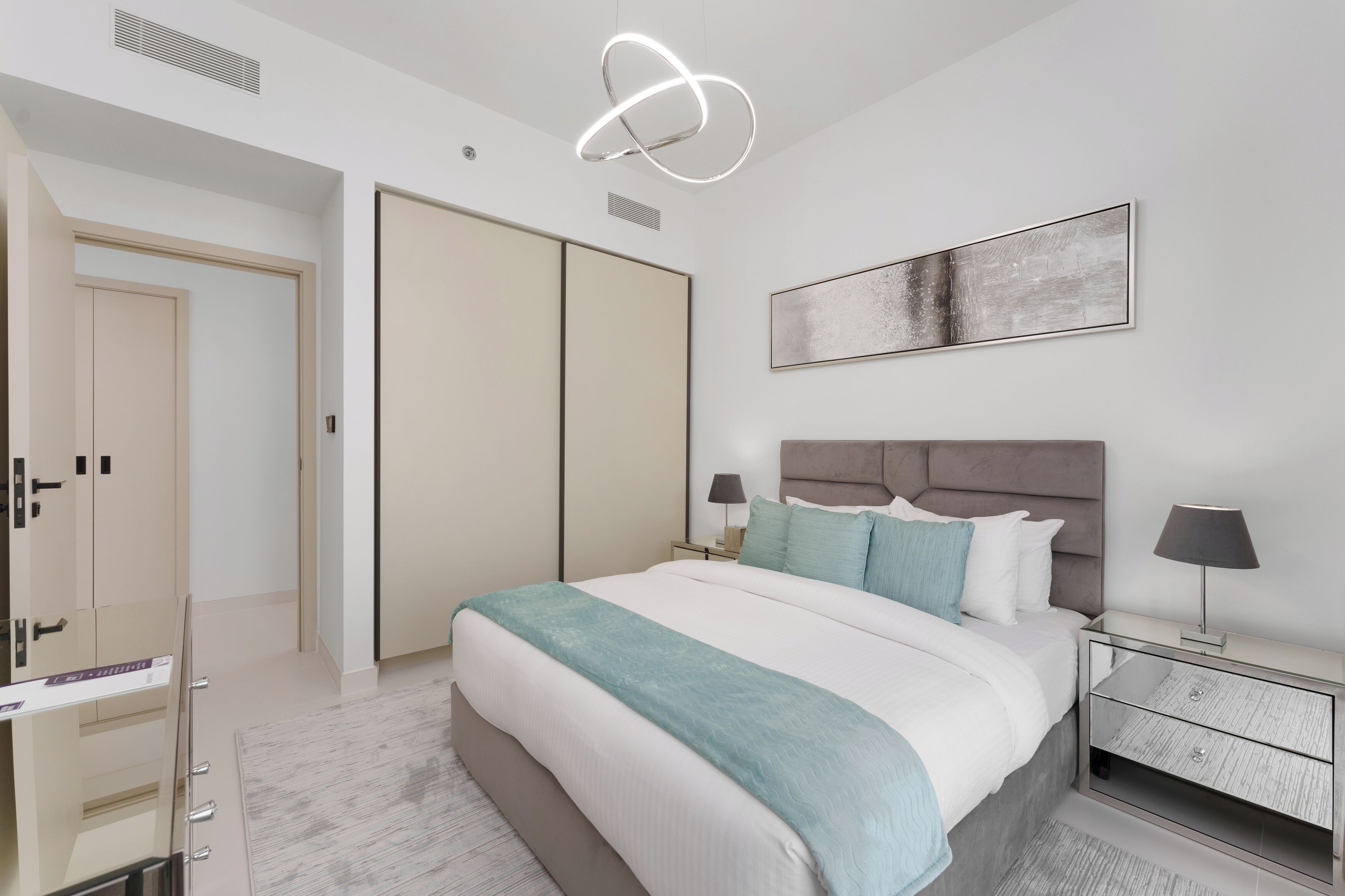 A modern bedroom with a large bed, a bedside table with lamps, a wardrobe, and a ceiling light fixture.