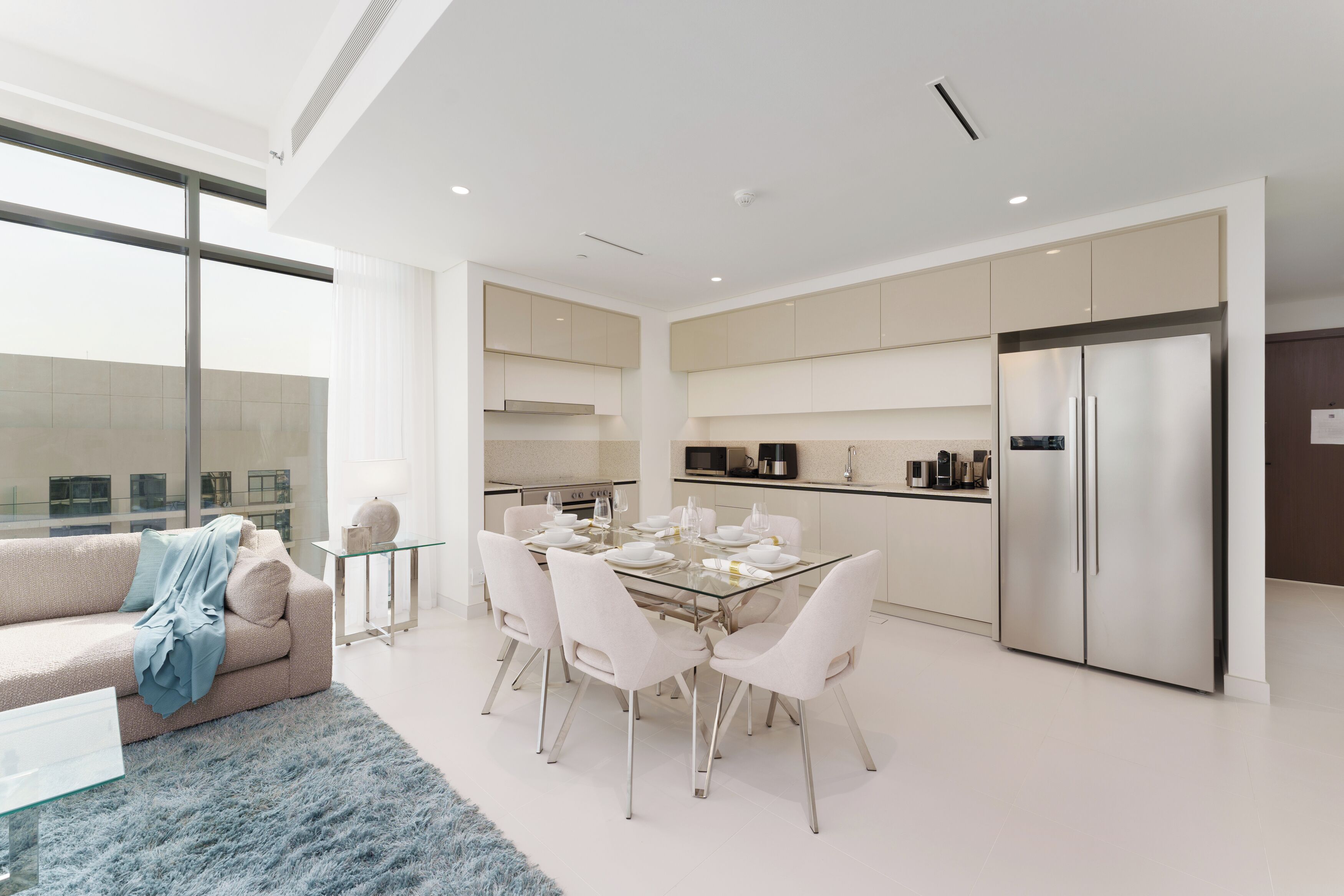 A modern kitchen with white cabinets, a stainless steel refrigerator, and a dining area with a glass table and white chairs.