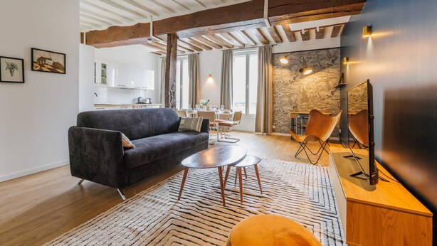 A modern living room with a dark sofa, a round coffee table, and a patterned rug. There is a dining area with a table and chairs, and a kitchen in the background. - Résidence Paris SEVRES (Paris)