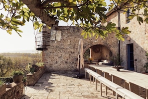An outdoor patio with stone flooring, a long wooden table, benches, and potted plants.