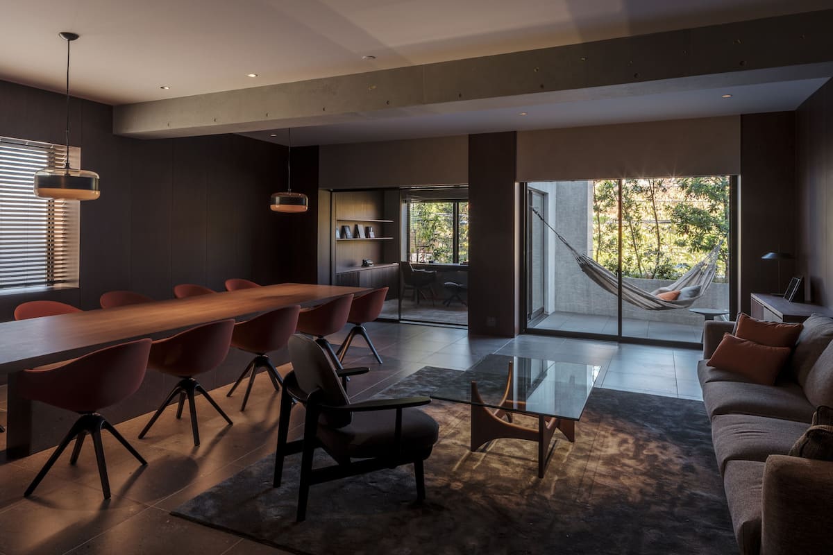 Un intérieur moderne avec une longue table à manger, des chaises rouges, une table basse en verre et un canapé. Un hamac est installé près d’une grande fenêtre.