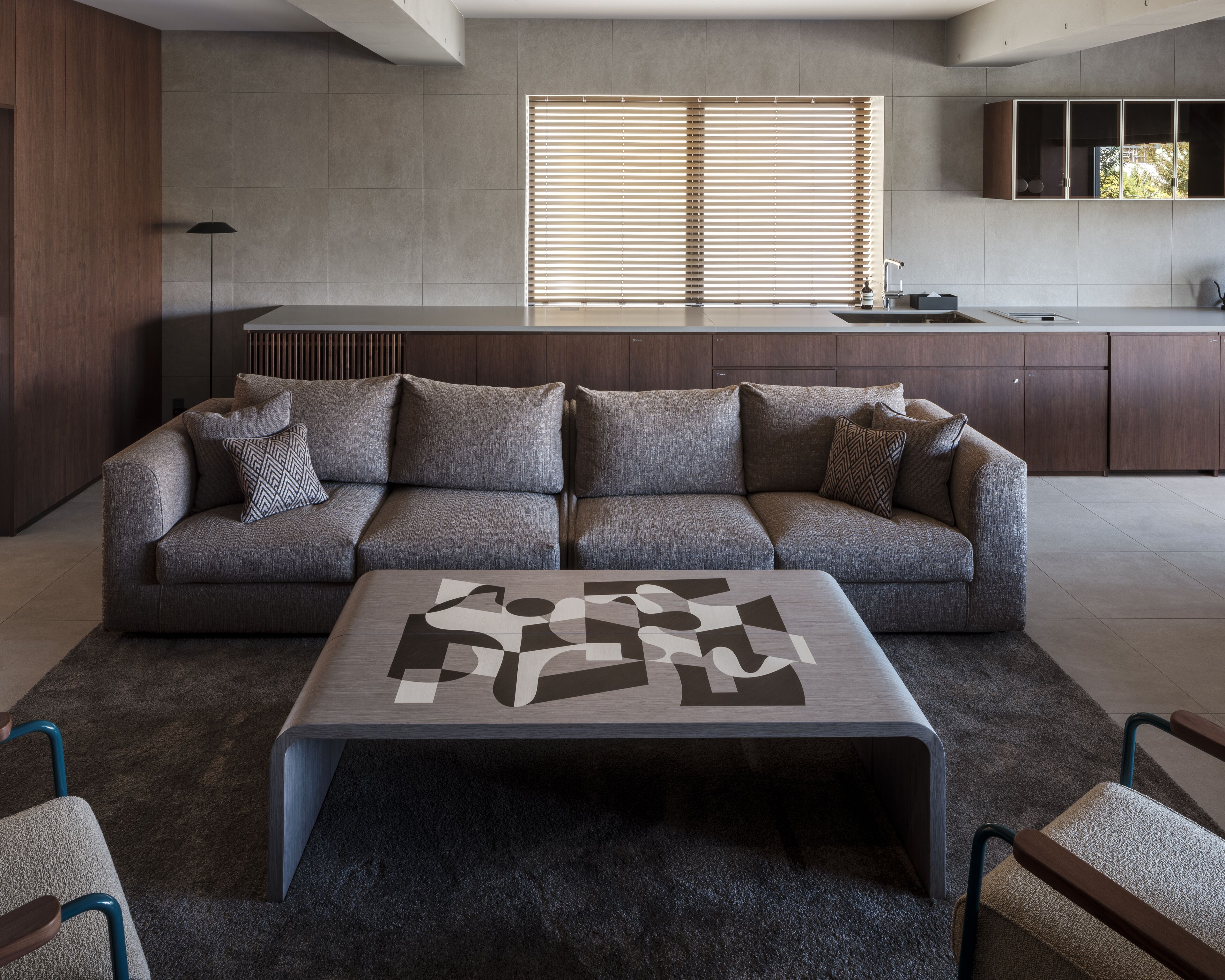 A modern living room with a grey sofa, a coffee table with a geometric design, and a large window with blinds.