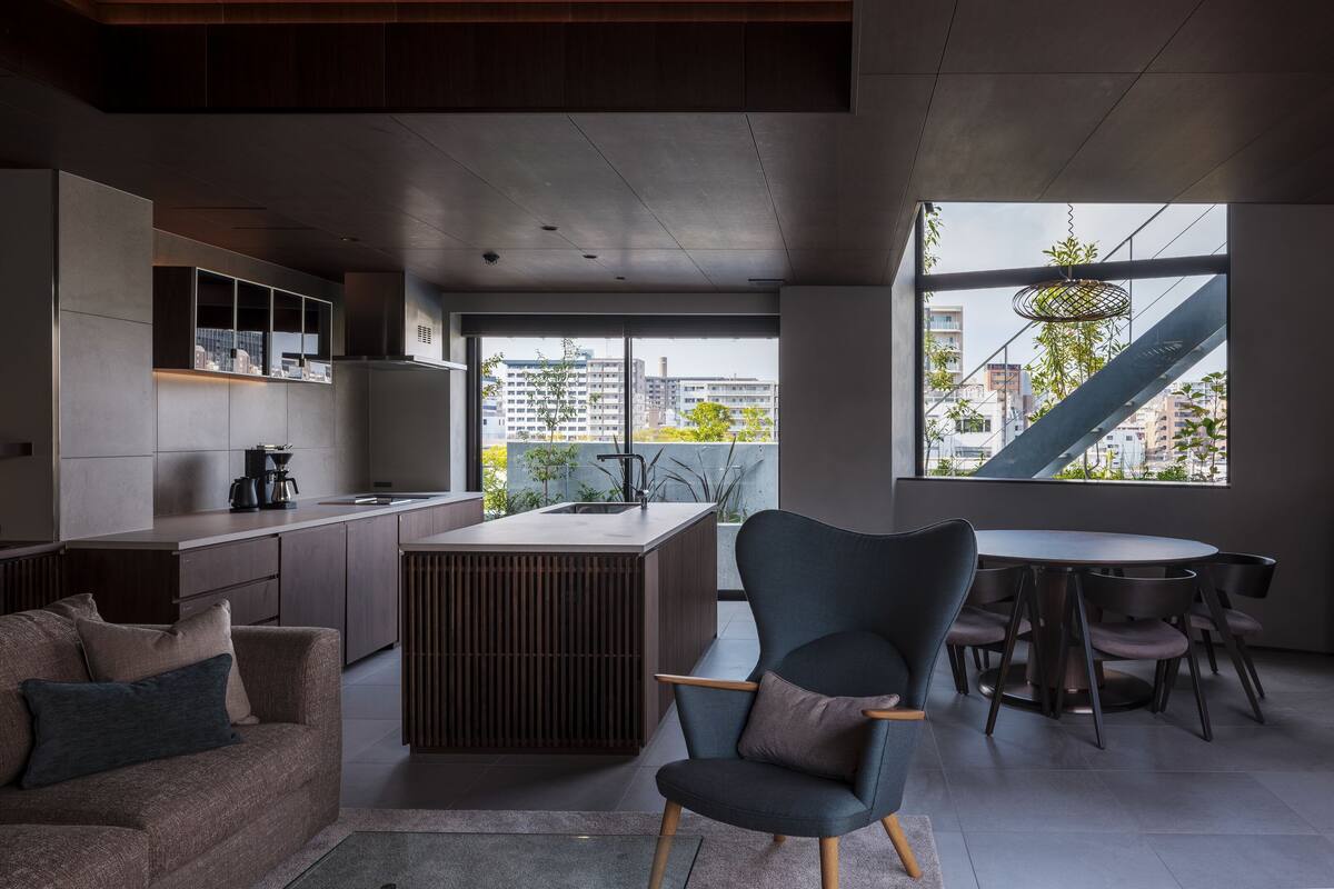 Une cuisine moderne dotée d’un îlot central, un coin repas avec une table ronde et des chaises, un canapé avec des coussins et une vue sur la ville grâce à de grandes fenêtres.