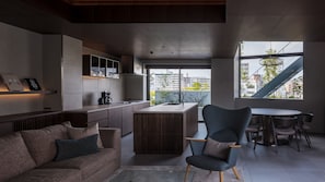 Une cuisine moderne dotée d’un îlot central, un coin repas avec une table ronde et des chaises, un canapé avec des coussins et une vue sur la ville grâce à de grandes fenêtres.