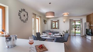 Ein moderner Essbereich mit einem weißen Tisch, einem Weinglas und Erdbeeren. Im Hintergrund ist eine Küchenzeile mit Fernseher und Esstisch zu sehen.