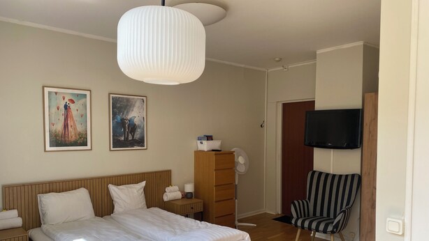A bedroom with a bed, a wooden nightstand, a bedside table with a lamp, a chair, a TV mounted on the wall, and two framed pictures on the wall. - På Hörnet (Karlskrona)