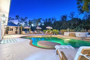 A swimming pool with lounge chairs and umbrellas, surrounded by trees and a building with a porch. - Hermosa Hacienda Whole House (Fort Myers)