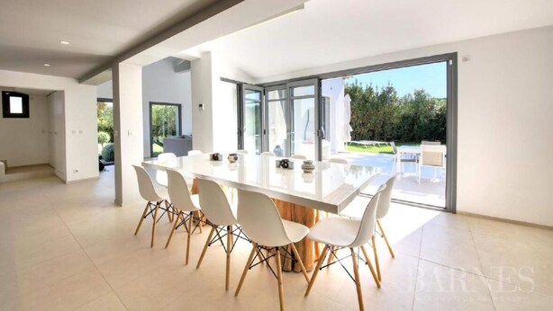 A modern dining area with a large white table and white chairs, a glass door leading to an outdoor patio, and a minimalist interior design. - Villa Contemporaine Neuve Plage a Pieds Piscine Chauffée (Saint-Tropez)