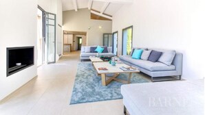 A spacious living room with a large sofa, a coffee table, and a TV mounted on the wall. - Villa Contemporaine Neuve Plage a Pieds Piscine Chauffée (Saint-Tropez)