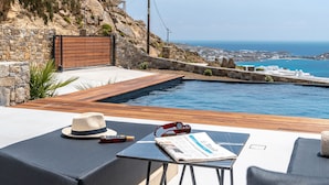 A poolside area with a wooden deck, lounge chairs, and a view of the sea. - Elit Villa Psarou Mykonos (Mykonos)