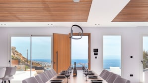 A modern dining room with a long wooden table set for a meal, large windows with a view of the ocean, and a wooden ceiling. - Elit Villa Psarou Mykonos (Mykonos)