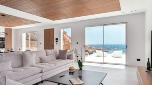 A modern living room with a large sectional sofa, a coffee table, and a view of the ocean through large sliding doors. - Elit Villa Psarou Mykonos (Mykonos)