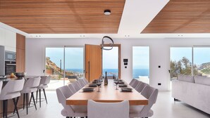 A modern dining area with a long wooden table, white chairs, and a view of the ocean. - Elit Villa Psarou Mykonos (Mykonos)