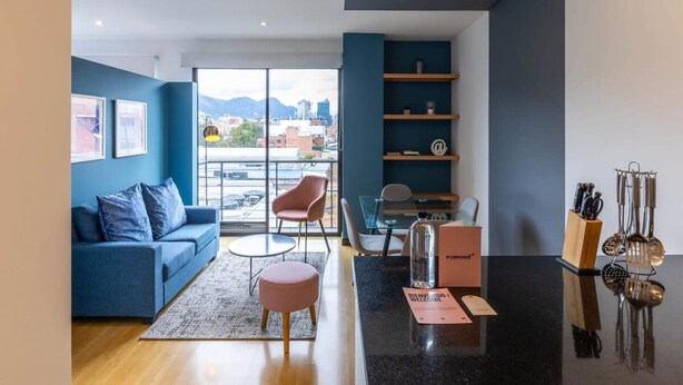 A modern living room with a blue sofa, a round coffee table, and a pink ottoman. There is a glass dining table with chairs, a bookshelf, and a kitchen area with utensils. - w Vibrant 1BR in Chico (Bogotá)