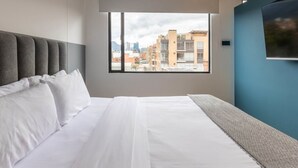 A hotel room with a large bed, a city view through a window, and a blue accent wall. - w Vibrant 1BR in Chico (Bogotá)