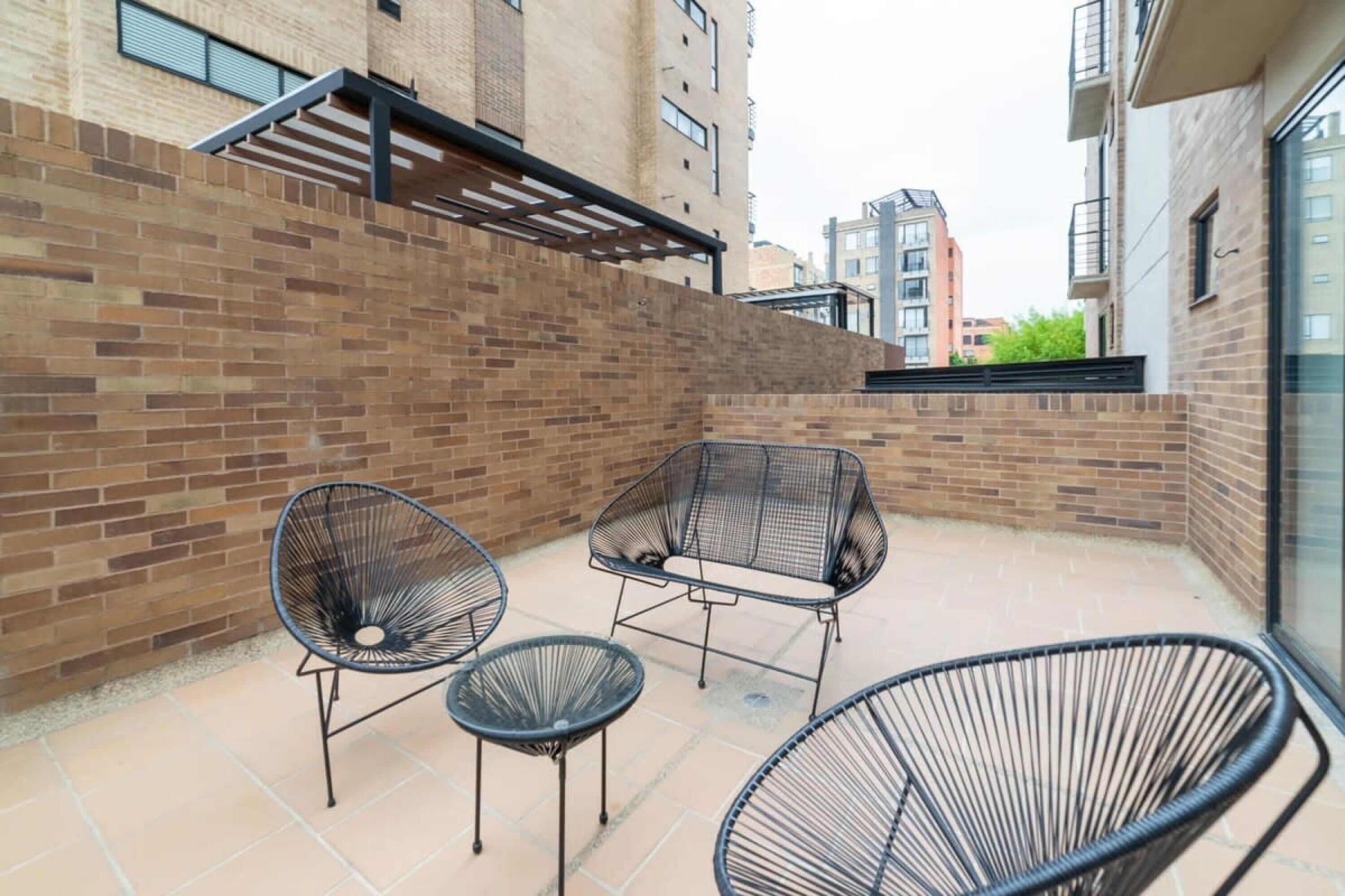 A courtyard with brick walls, a pergola, and three black wireframe chairs.