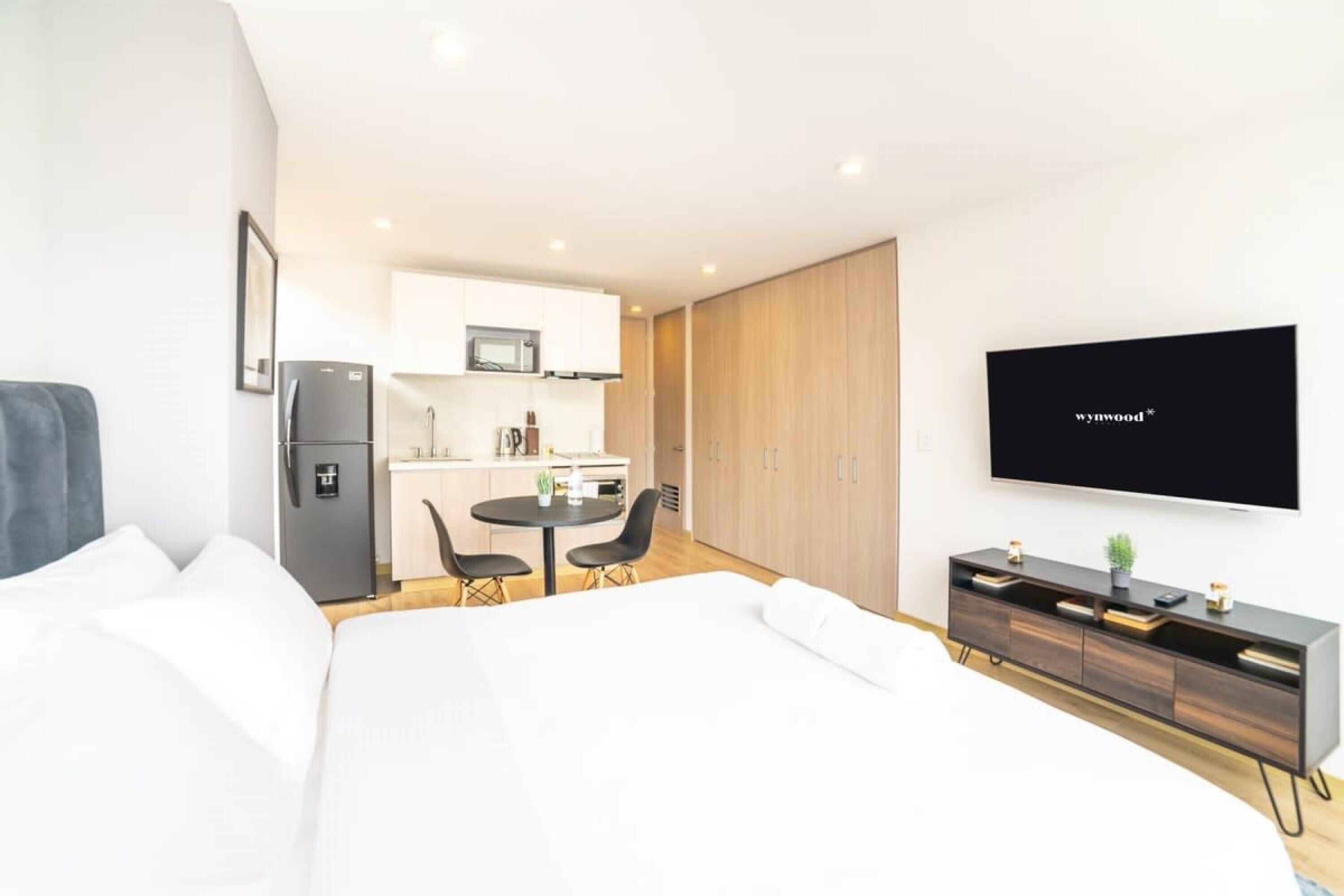 A modern hotel room with a bed, a small kitchenette, a dining table, and a TV mounted on the wall.