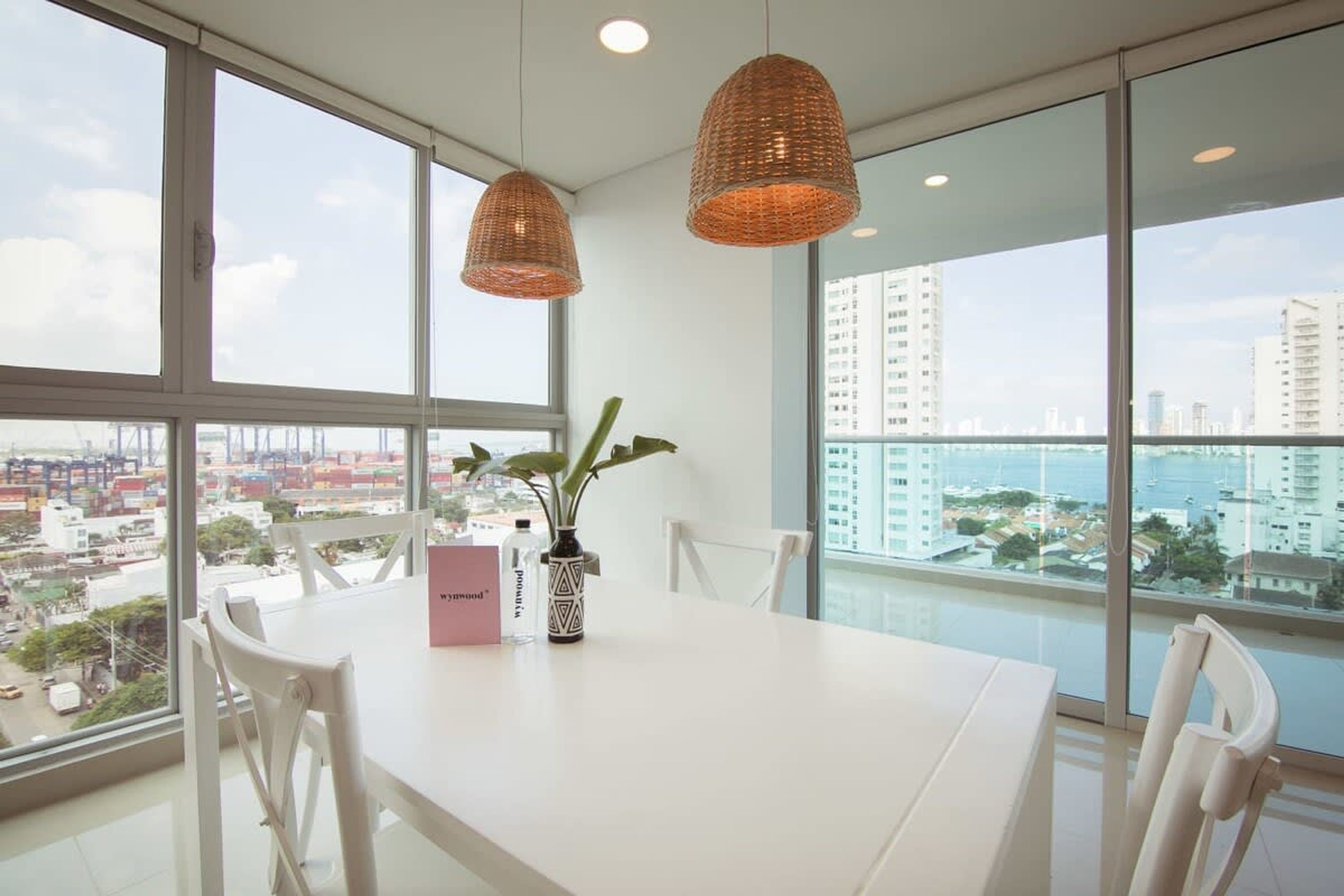 Uma sala de jantar moderna com mesa e cadeiras brancas, e vista para a cidade através de amplas janelas.