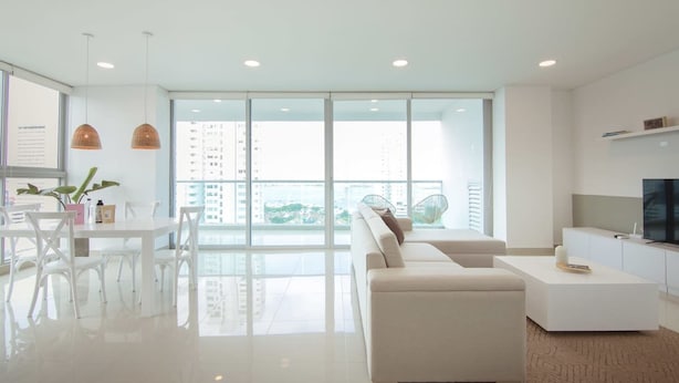 A modern living room with a white sofa, a dining table with chairs, a TV, and a balcony with a view of buildings. - w Modern 2BR w AC in Paradisiac Cartagena (Cartagena)