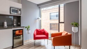 Une cuisine moderne équipée d’un four à micro-ondes, d’un petit réfrigérateur et d’un évier. On y trouve deux chaises, une rouge et une orange, ainsi que deux plantes en pot. La pièce est dotée de grandes fenêtres donnant sur un immeuble en briques.
