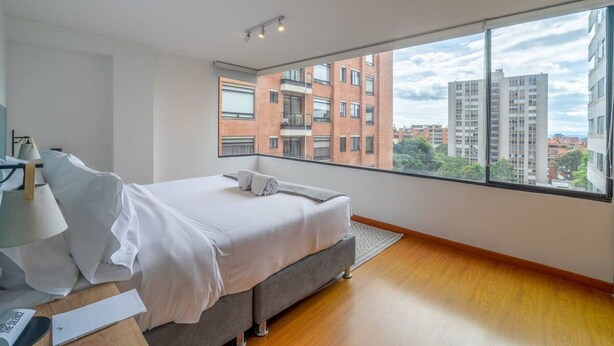 A modern bedroom with a large window, a bed, a bedside table, and a lamp.