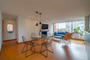 A modern living room with a round glass table, black wireframe chairs, a blue sofa, and a large window offering a city view. - w Centric 2BR In Cabrera (Bogotá)