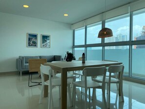 A modern dining area with a wooden table and white chairs, large windows with a view of buildings, a grey sofa, and two framed pictures on the wall. - w Spectacular 3BR w AC in Cartagena (Cartagena)