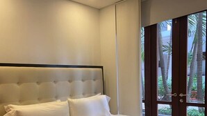 Une chambre d’hôtel avec un grand lit, une tête de lit et une vue sur des palmiers, visible à travers une porte.