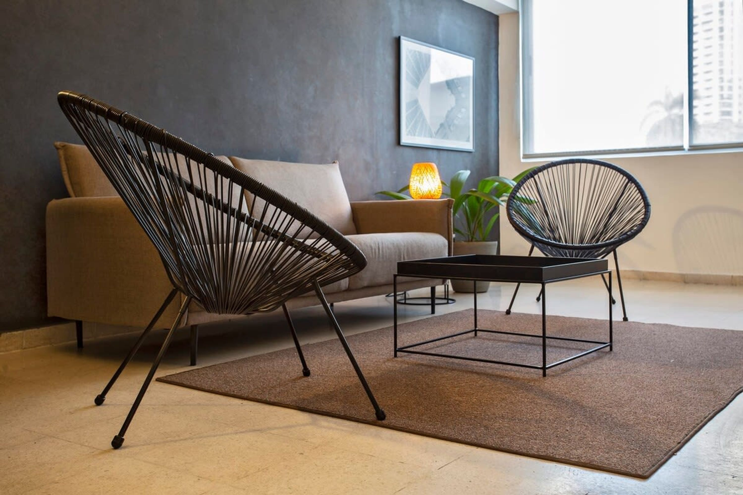 A modern living room with a sofa, two wireframe chairs, a small table, a lamp, and a plant.