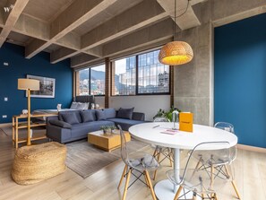 A modern living room with a blue wall, a gray sofa, a round dining table with clear chairs, and a hanging light fixture.
