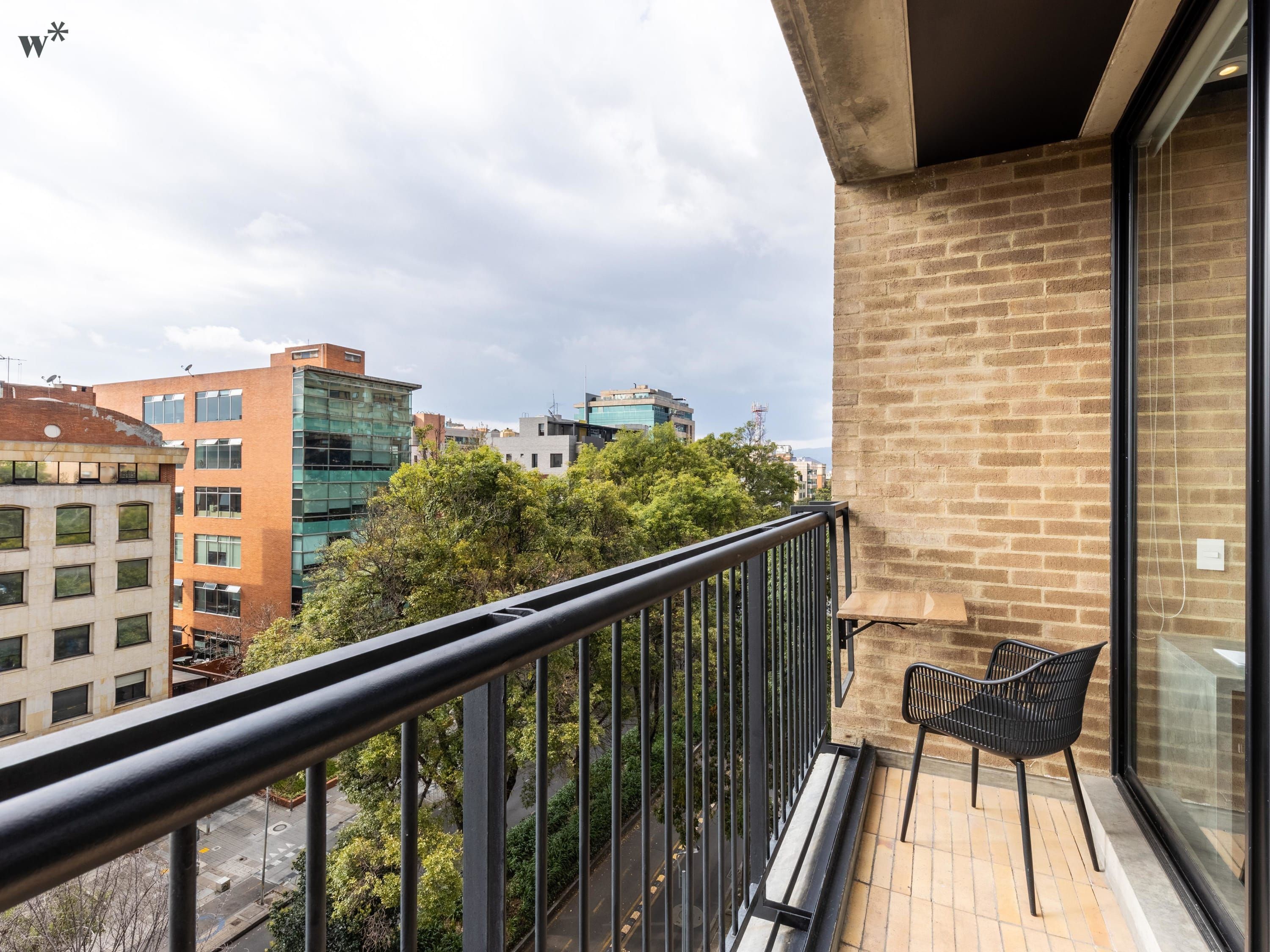 Un balcon doté d’une balustrade en métal noir, d’une chaise noire et offrant une vue sur un paysage urbain avec des bâtiments et des arbres.