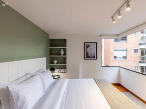 A modern bedroom with a bed, a bookshelf, and a window with a city view. - w Homey 2BR in Cabrera (Bogotá)