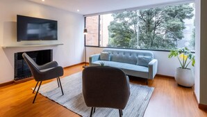 Uma sala de estar moderna com televisão de tela plana na parede, um sofá, duas poltronas, lareira e uma grande janela com vista para árvores.