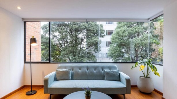 Uma sala de estar moderna com um sofá cinzento, uma mesa de centro redonda e uma janela ampla com vista para árvores e edifícios.