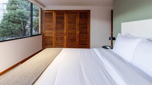 Quarto com cama grande, guarda-roupa de madeira, mesa de cabeceira e vista para árvores através de uma janela.