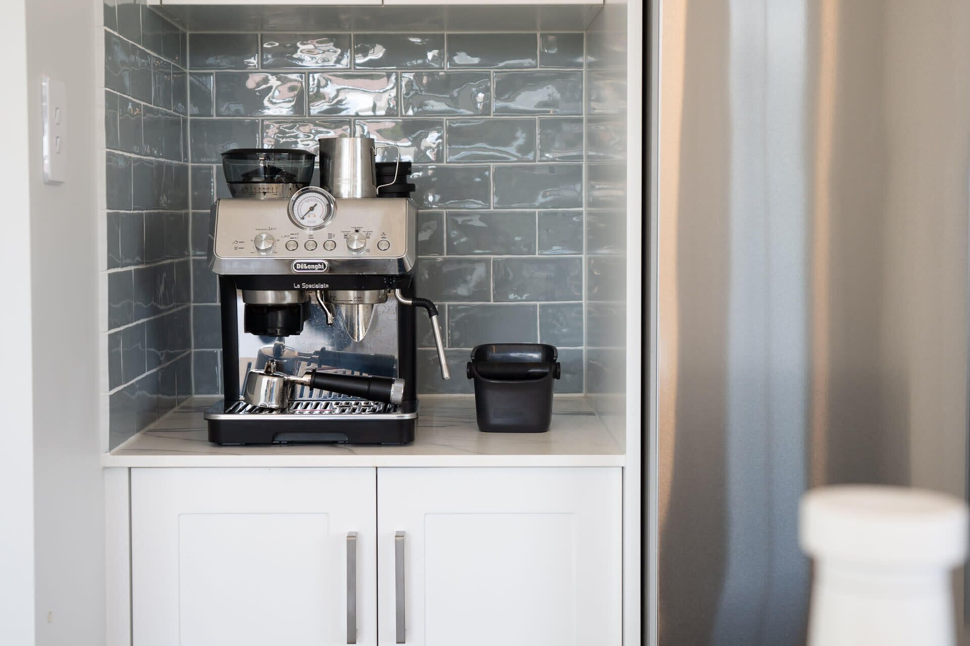 Máquina de café em cozinha com painel de azulejos e armários brancos.