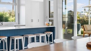 A modern kitchen with a large window, white cabinets, and a dining area with stools. - 6 Beach Rd Goolwa South (Goolwa South)
