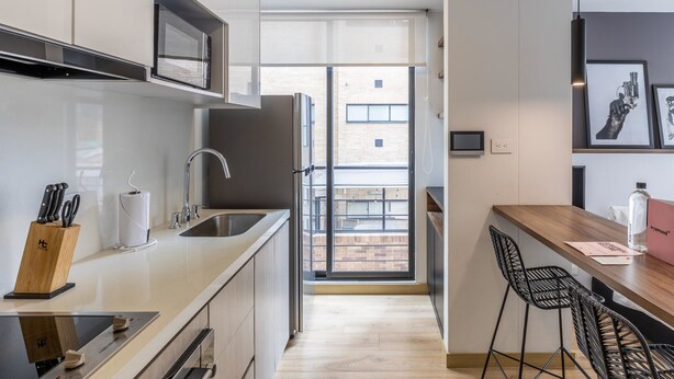 A modern kitchen with a built-in microwave, stainless steel sink, and a black range hood. There is a wooden cutting board with knives, a roll of paper towels, and a glass bottle on the counter. The kitchen leads to a balcony with a view of a brick building. - w Splendid Loft in Santa Ana (Bogotá)