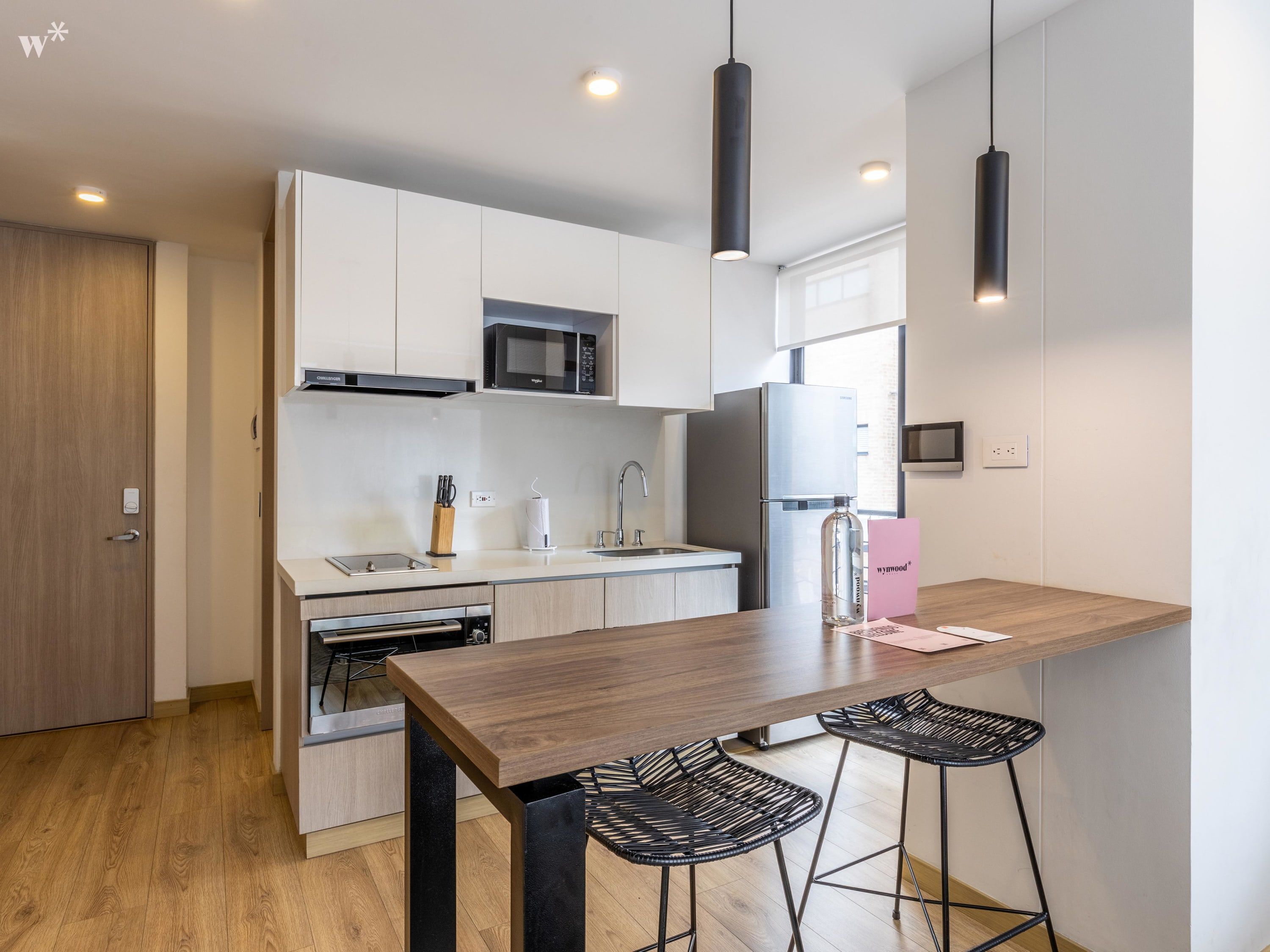 A modern kitchen with white cabinets, a stainless steel refrigerator, a microwave, and a sink. There are two black bar stools and a wooden table.