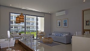 A modern living room with a sofa, chairs, a coffee table, and a dining area with a table and chairs. There is a large window providing a view of a building outside.